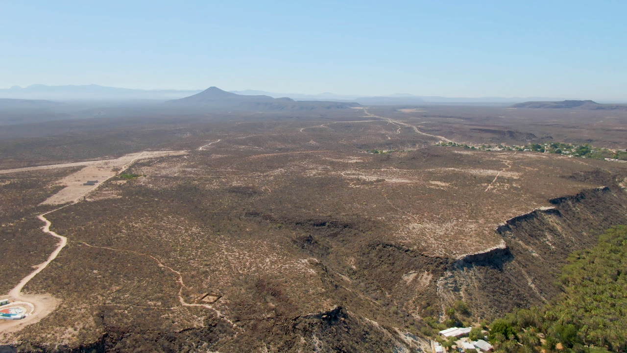 Wide aerial view of arid terrain, winding road, rocky ridge, green trees, and a distant mountain.