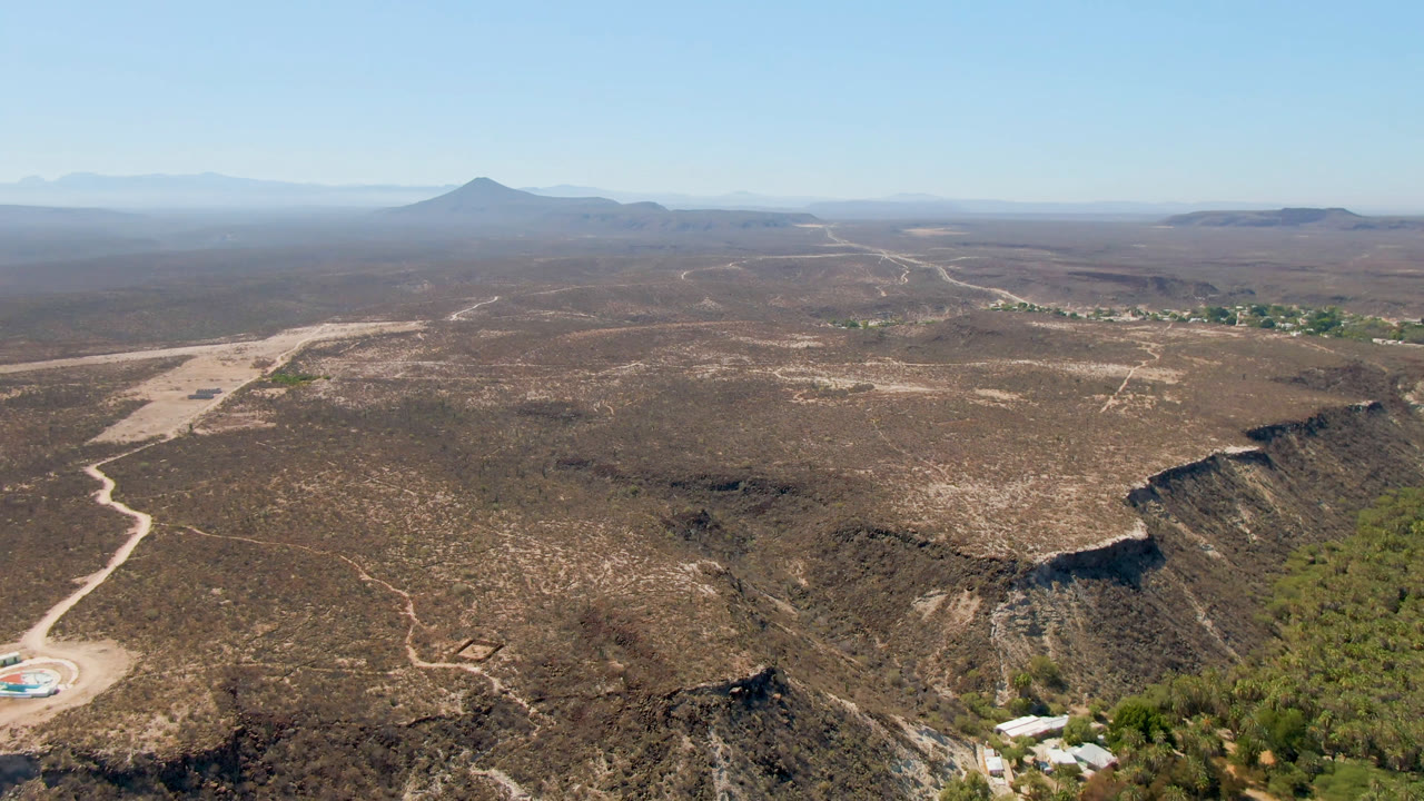 Early aerial view in San Ignacio, Baja California Sur, with the winding road more centered across the desert flats, a green settlement strip in the distance, a mountain on the horizon, and no visible people.