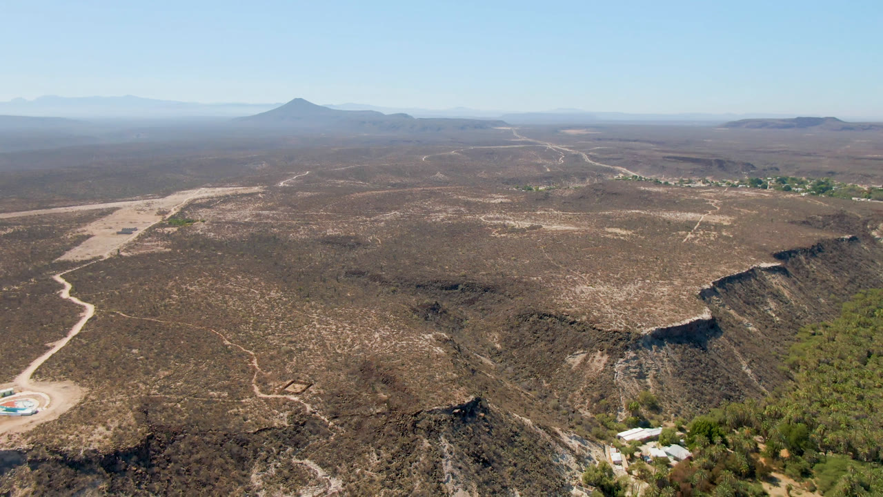 High aerial view of dry desert, rocky ridge, trees, road, and distant mountain under clear sky.