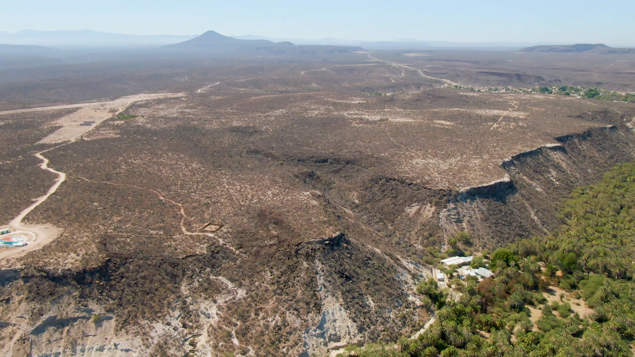 Wide aerial view of desert plateau, dirt road, green tree corridor, and distant mountains.