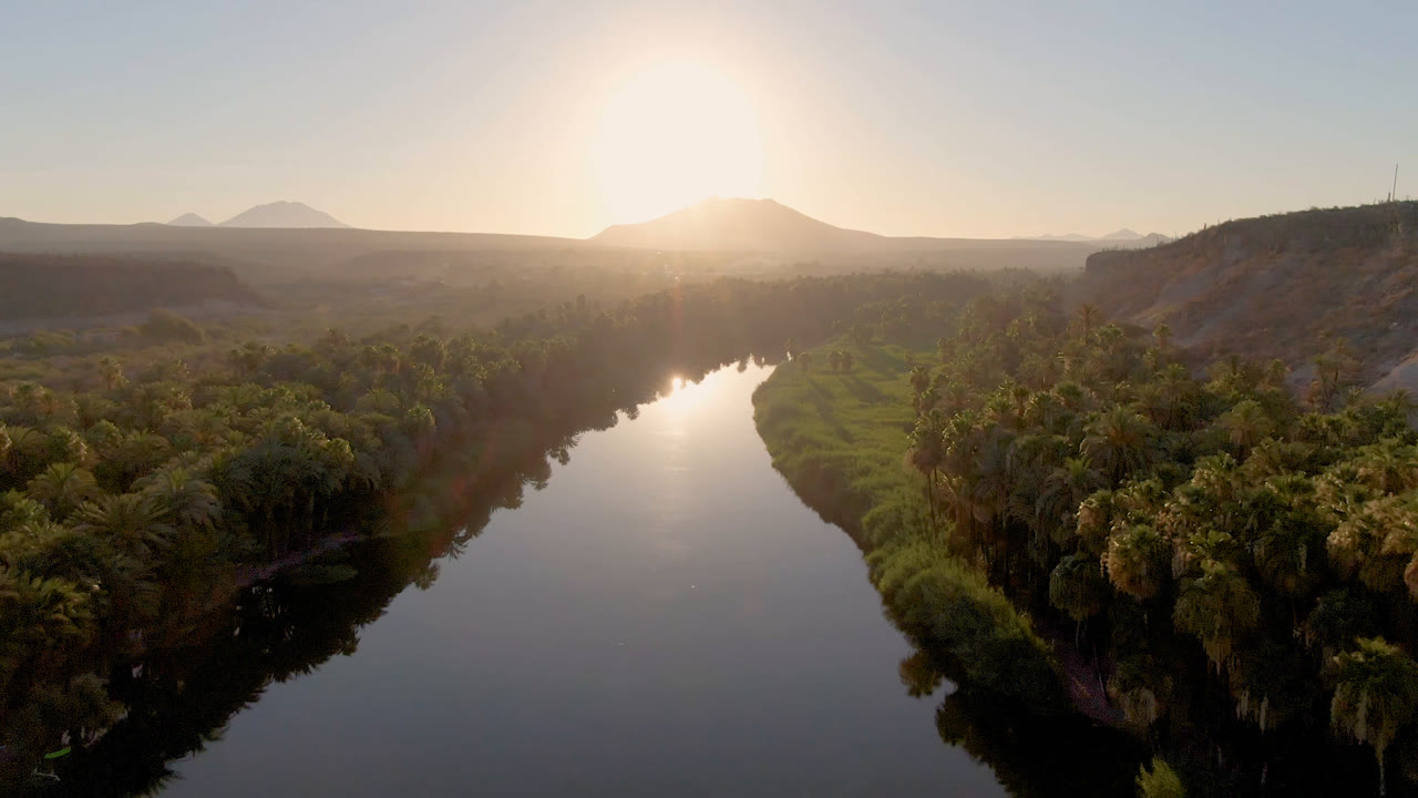 Opening aerial frame over San Ignacio, Baja California Sur, Mexico, showing a reflective river bordered by palms, dry ridges, distant mountains, bright low sun, and no visible people.