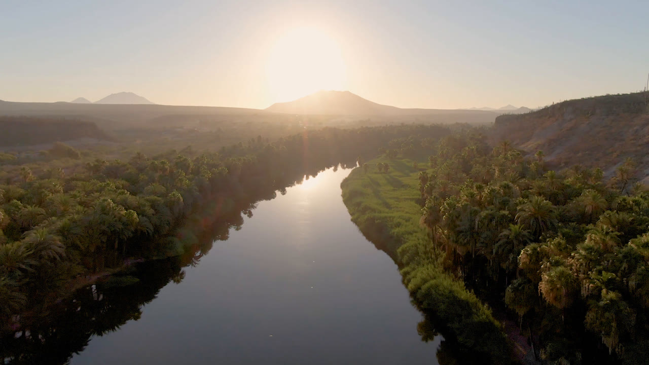 Aerial frame over San Ignacio, Baja California Sur, Mexico, with the river reflecting the sun, palms filling the lower right, a dry ridge on the right side, distant mountains, and no visible people.