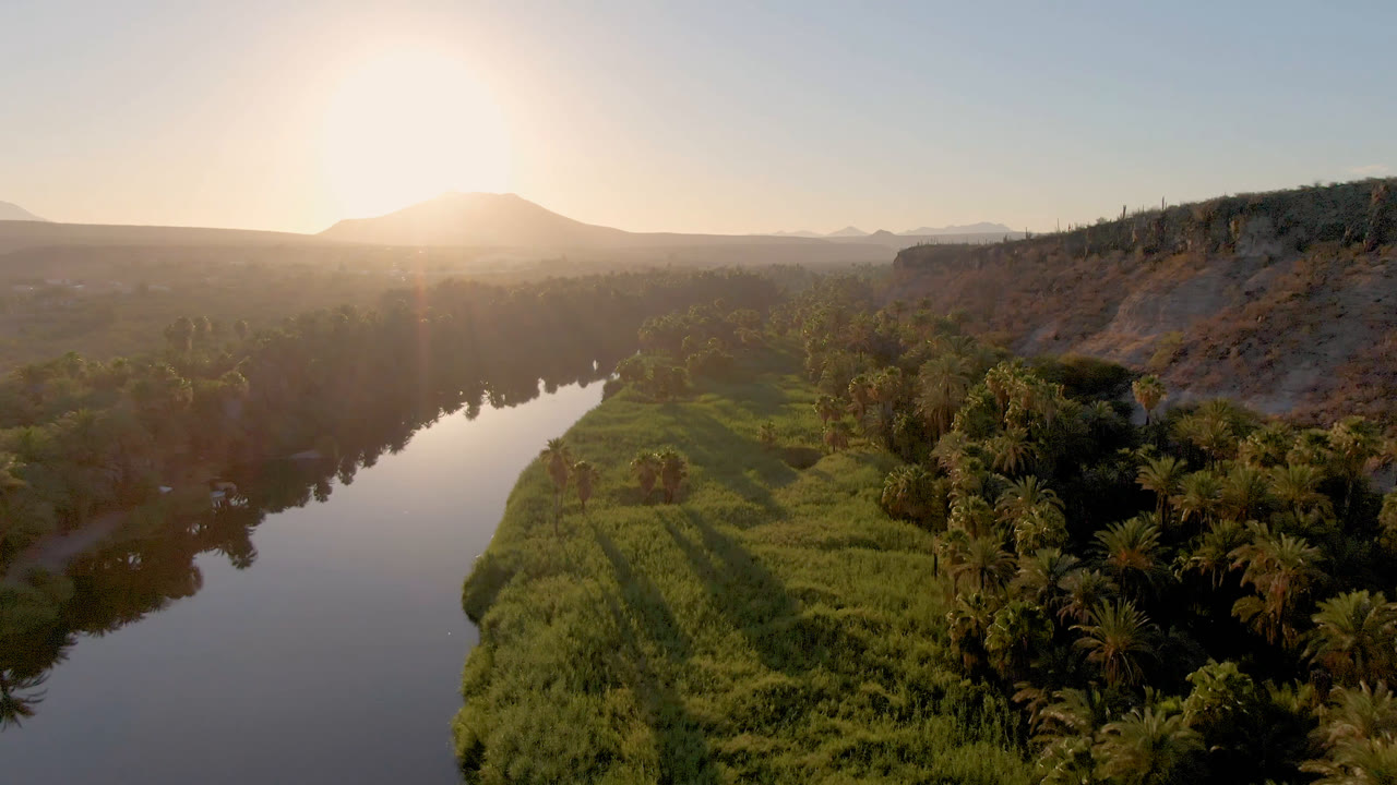 River, green shoreline, palms, ridge, and low sun over distant mountains.