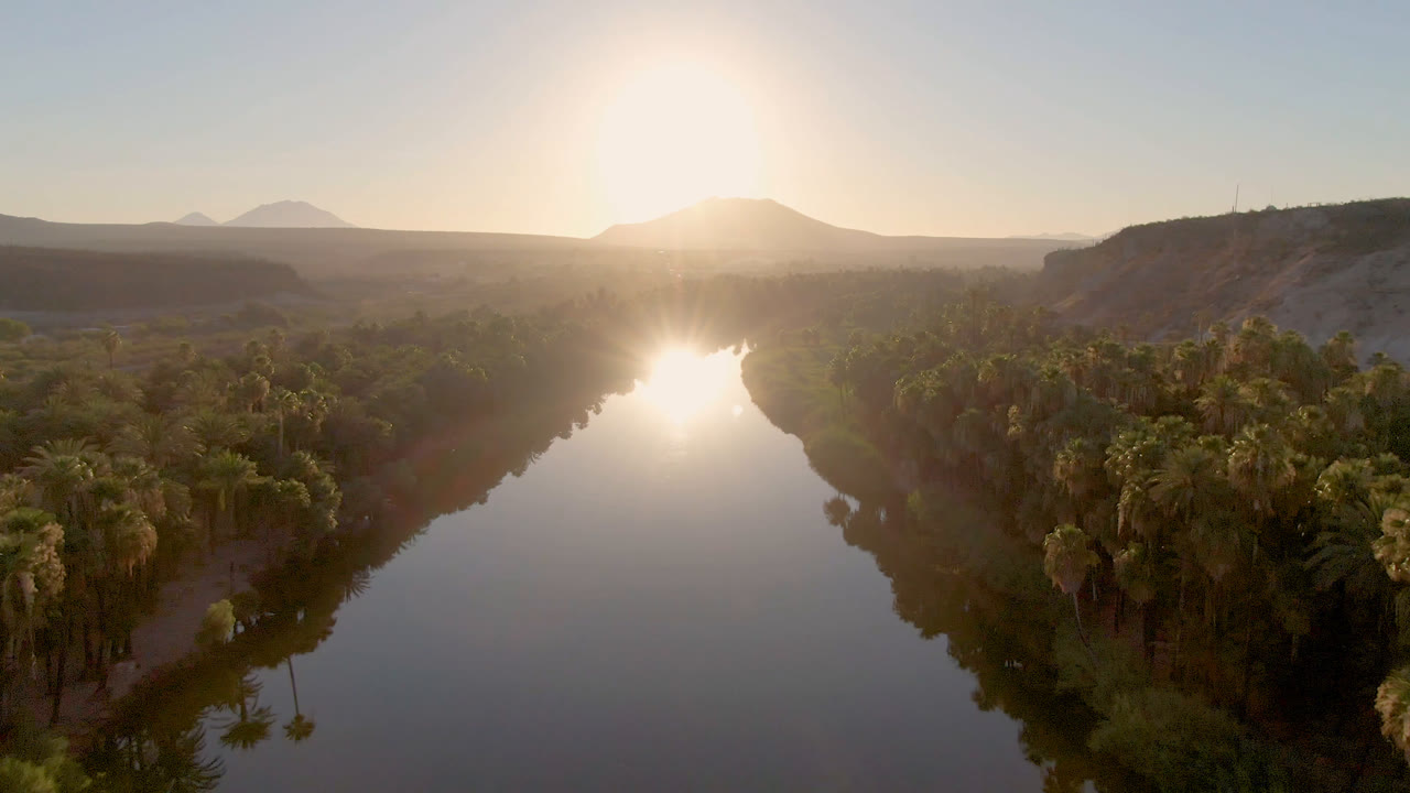 Aerial view over a calm river in San Ignacio, Baja California Sur, Mexico, with palm-lined banks, dry hills, distant mountains, low sun glare on the water, and no visible people.