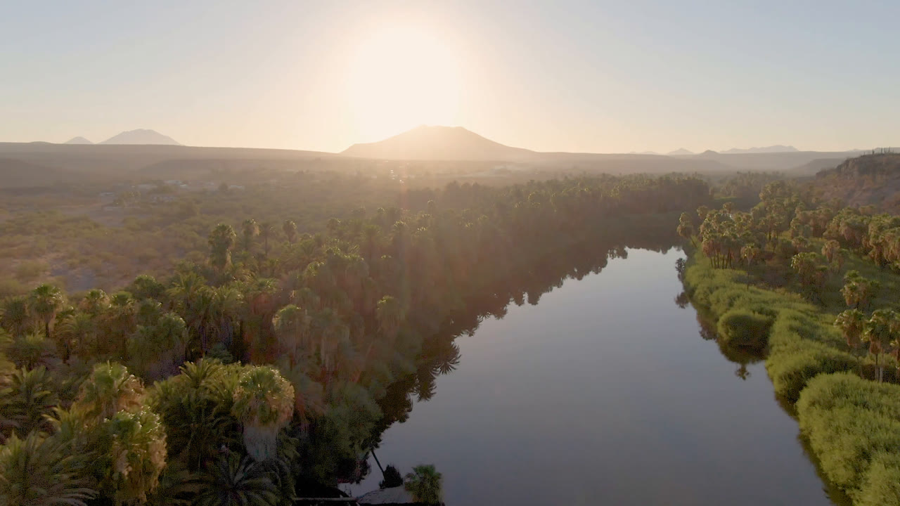 A later aerial view in San Ignacio, Baja California Sur shows the river widening through green vegetation, with a hazy mountain ridge, pale sky, and no visible people.
