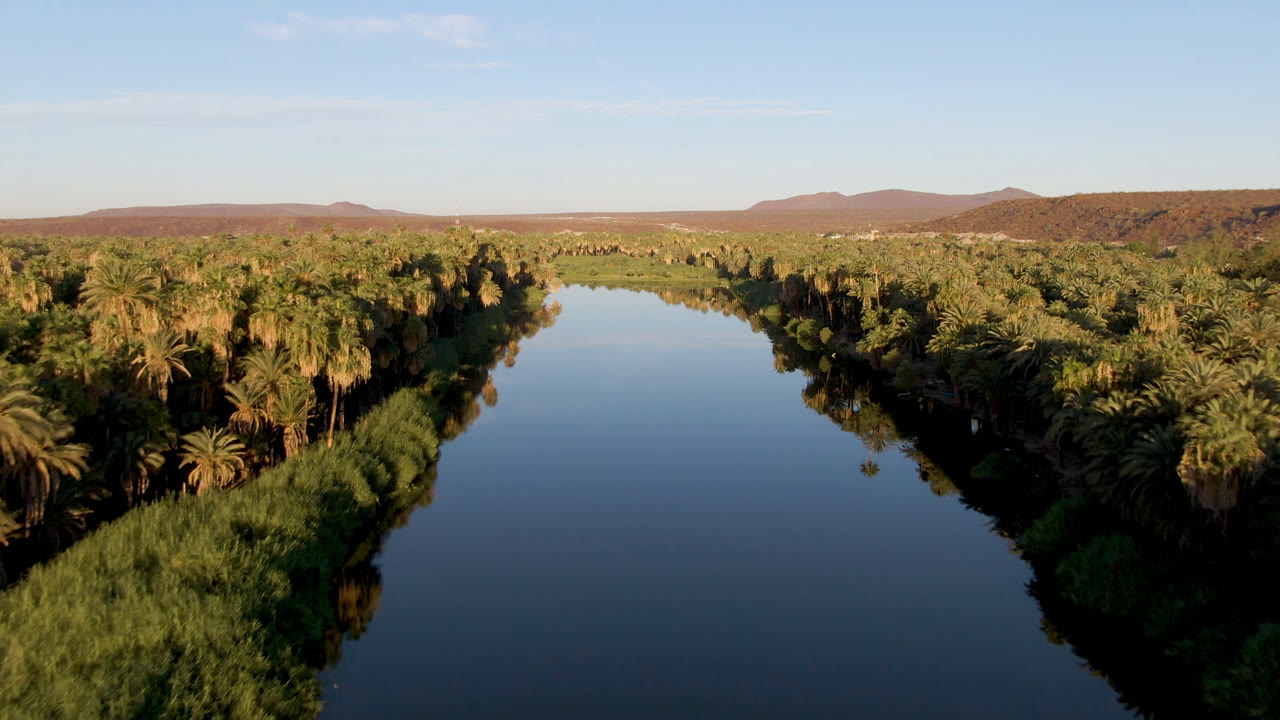 A slightly later aerial view in San Ignacio shows the river running between shadowed palm banks, with blue sky reflected in the water and no visible people.