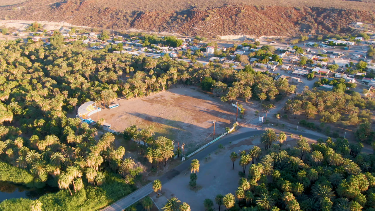 Wide aerial of green palms, a dirt clearing, small buildings, and mountains.