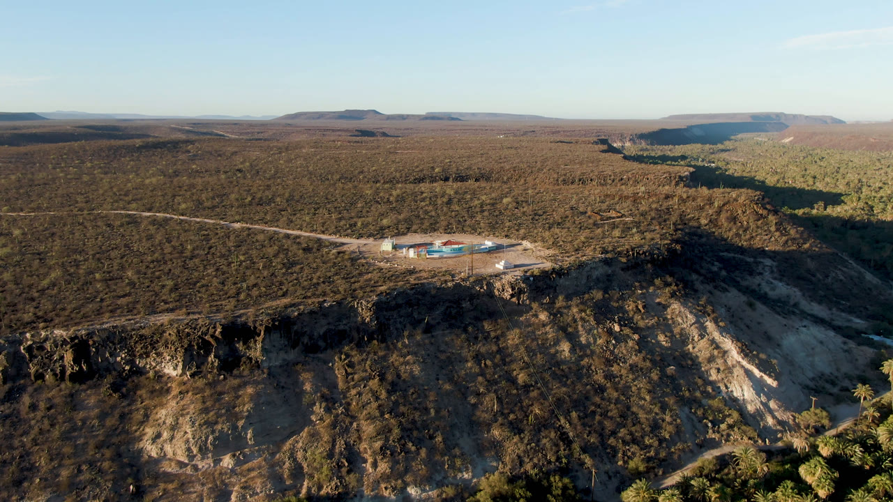 Wide aerial frame over San Ignacio, Baja California Sur, showing a cliff edge in the foreground, a low building complex with blue pools in the midground, dry plateau and mesas beyond, and no visible people.