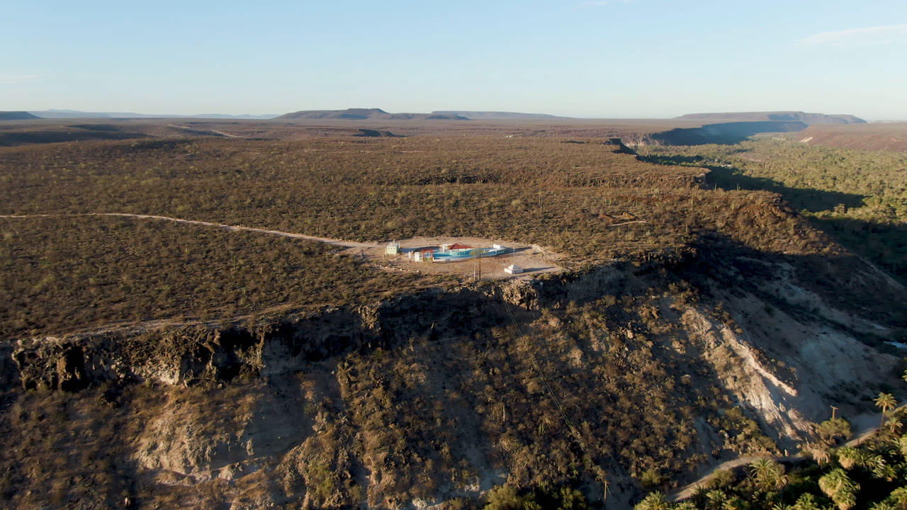 Aerial view in San Ignacio, Baja California Sur, with the cliff face filling the lower frame, the building complex and blue pools set back from the edge, distant mesas on the horizon, and no visible people.