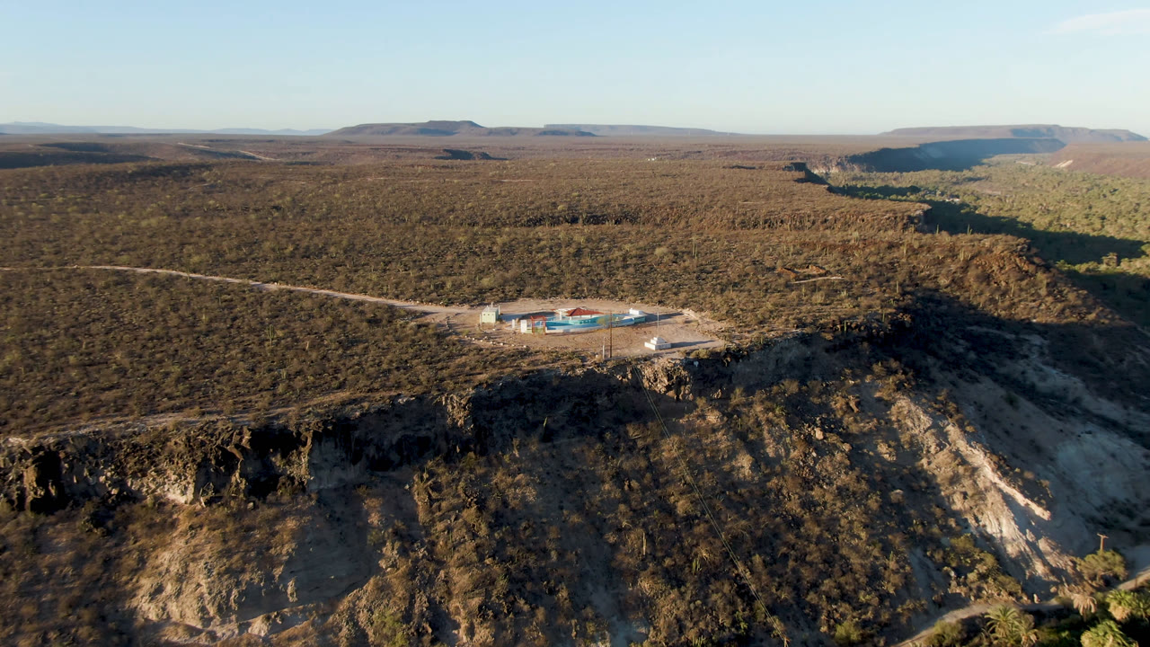 High drone-style composition over San Ignacio, Baja California Sur, showing a low compound with blue pools near a cliff, a green ribbon of vegetation in the canyon, dry brush across the plateau, and no visible people.