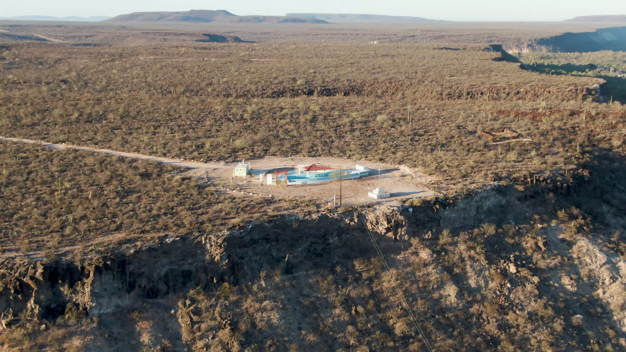 Aerial frame from San Ignacio, Baja California Sur, with the building complex centered against tan desert scrub, a pale road crossing the scene, distant mesas, and no visible people.