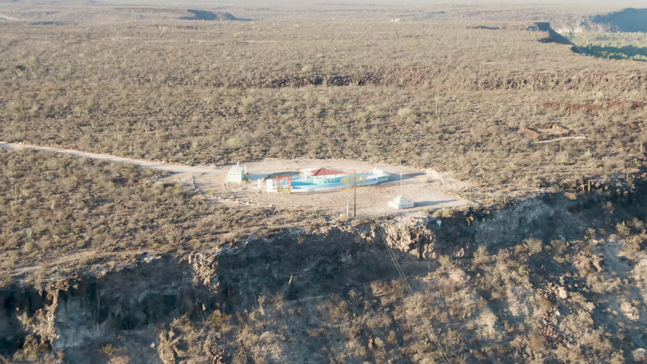 Wide aerial frame over San Ignacio, Baja California Sur, showing a pale building with blue and red areas beside a road on a rocky cliff, with scrub desert and no visible people.