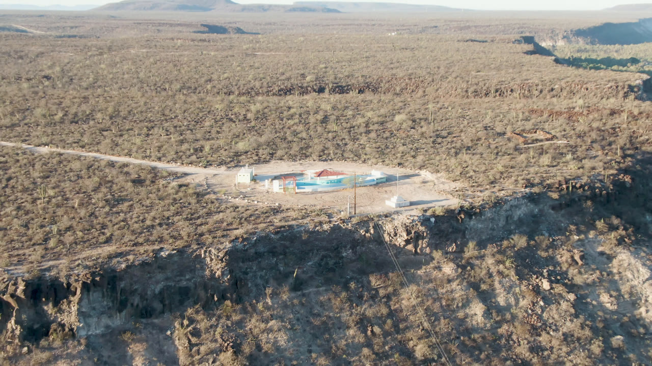 Aerial frame in San Ignacio, Baja California Sur, with the building slightly left of center, a road crossing dry terrain, dark cliff shadows in front, and no visible people.
