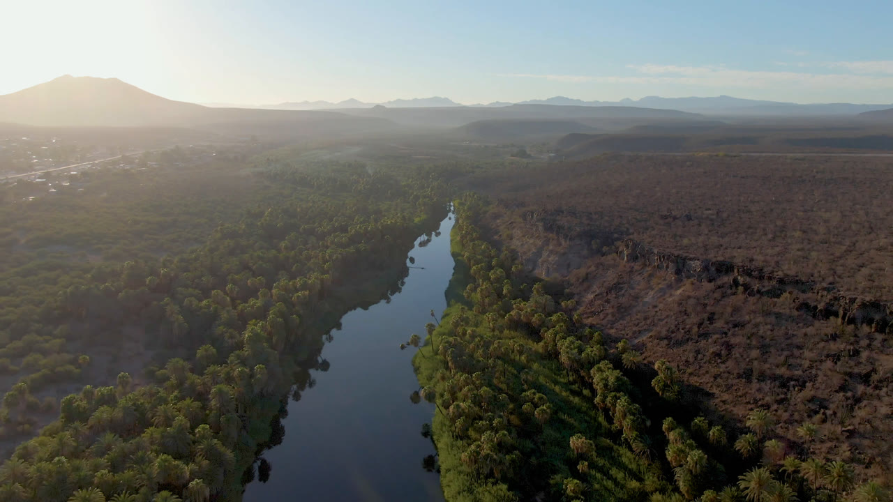 Aerial frame over San Ignacio, Baja California Sur, showing a dark river bordered by green trees, dry terrain to the right, hazy mountains beyond, and no visible people.