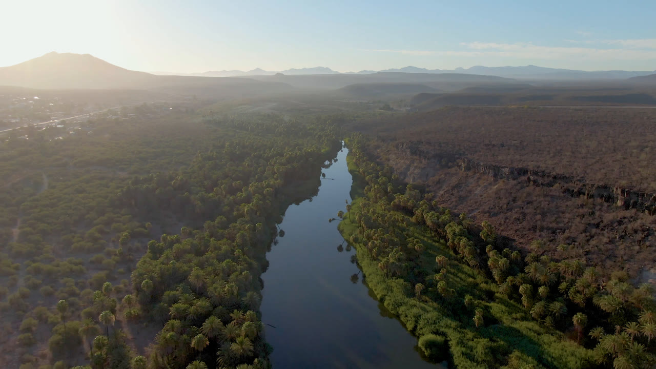 Aerial view in San Ignacio, Baja California Sur, Mexico, with a reflective river centered below, green trees along the banks, dry scrubland to the right, hazy mountains beyond, and no visible people.