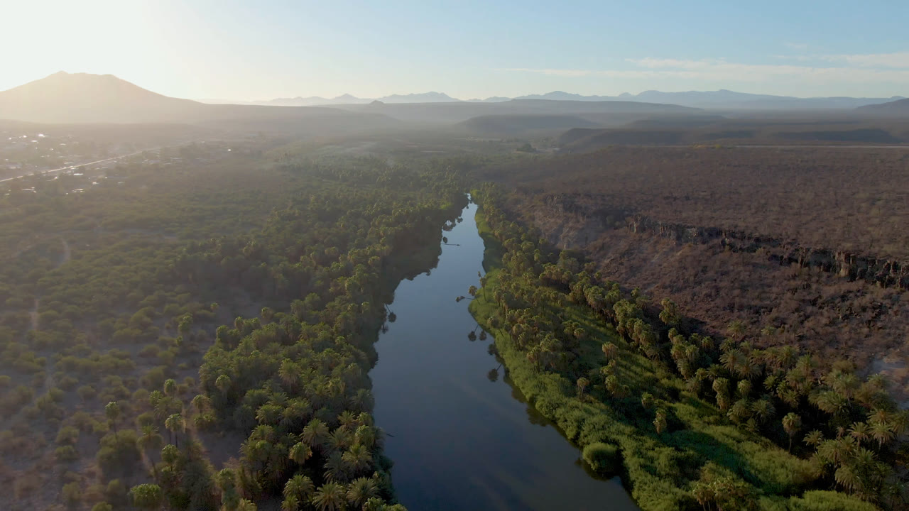 Drone view over San Ignacio with the narrow river running from the lower center toward the horizon, dense green bank vegetation, brown brushland on both sides, soft left-side sun flare, and no visible people.