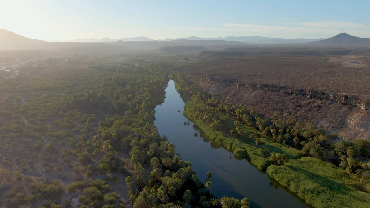Aerial frame over San Ignacio, Baja California Sur, showing a narrow river between dense green vegetation and dry scrubland, with distant mountains and no visible people.