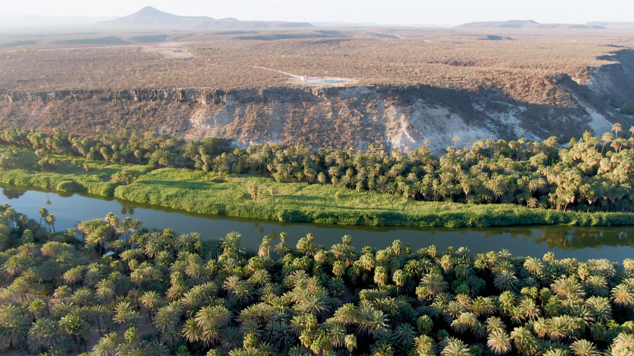 Opening aerial frame over San Ignacio, Baja California Sur, with a green river corridor below palms, pale desert bluffs beyond, distant hills on the horizon, and no visible people.