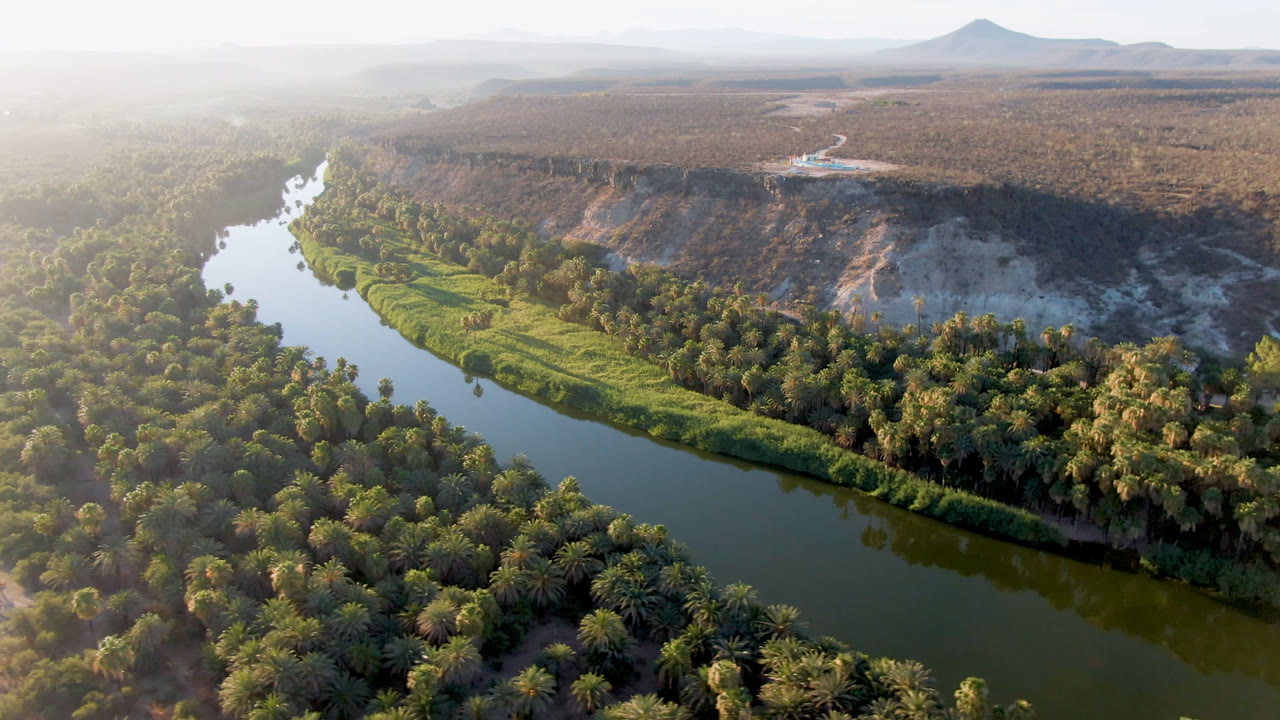 Second aerial frame over San Ignacio shows the river running along thick palms, with a sunlit green bank, pale eroded bluff, open scrubland, distant mountains, and no visible people.
