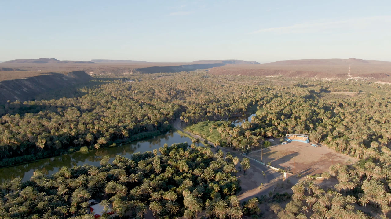 Wide aerial frame in San Ignacio, Baja California Sur, showing the river curving through palms, the cleared field shifted toward the lower right, distant ridges, and no visible people.