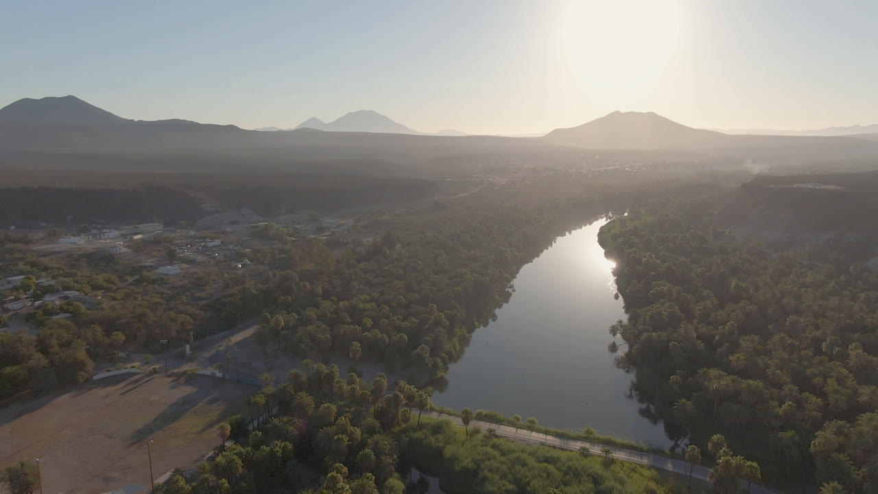 Opening aerial frame over San Ignacio, Baja California Sur, with a reflective river, green tree canopy, dry fields, distant mountains, hazy sky, and no visible people.