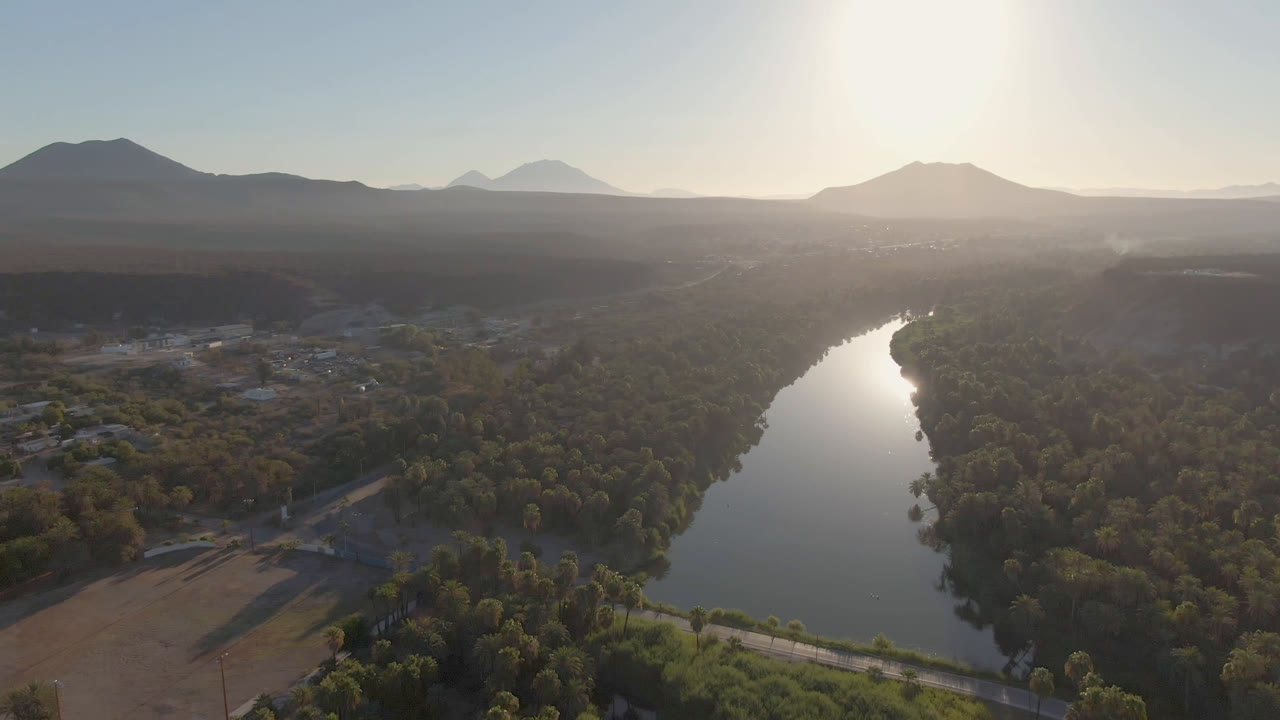 Second aerial frame in San Ignacio showing the river bend slightly more centered, with sun reflection on the water, tree-lined banks, fields, low mountains, and no visible people.
