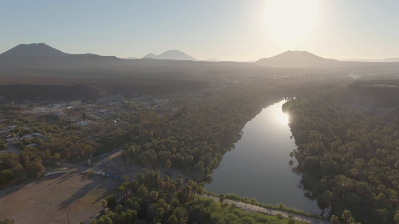Aerial landscape with a river, dense trees, open fields, and a bright mountain horizon.