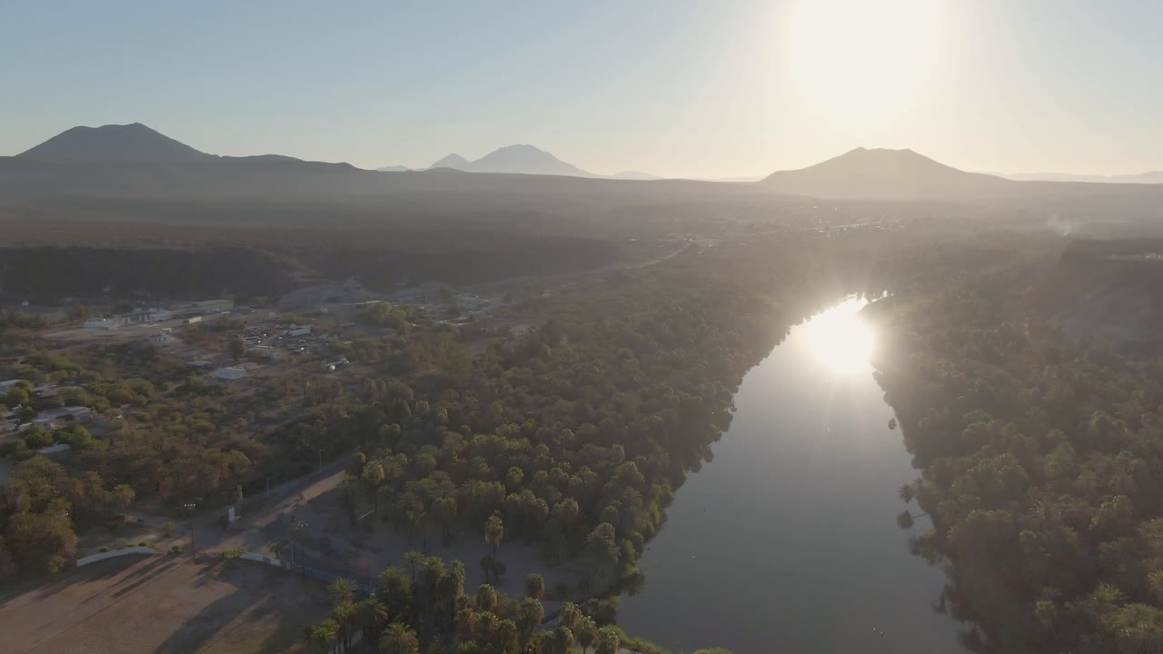 Aerial sunset view over a river in San Ignacio, Baja California Sur, Mexico, with palms and low buildings along the banks, distant mountains, and no visible people.