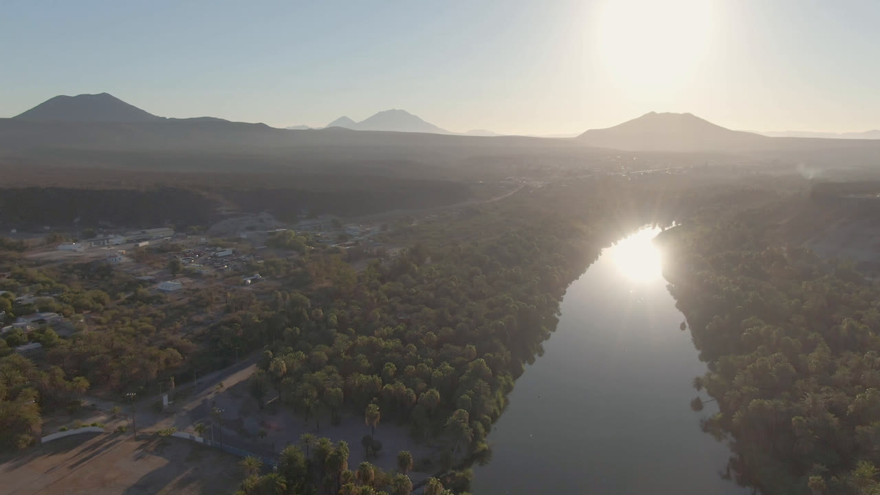 Wide aerial view in San Ignacio, Baja California Sur, Mexico, showing bright glare on the river, tree-lined banks, low settlement structures, distant mountains, and no visible people.