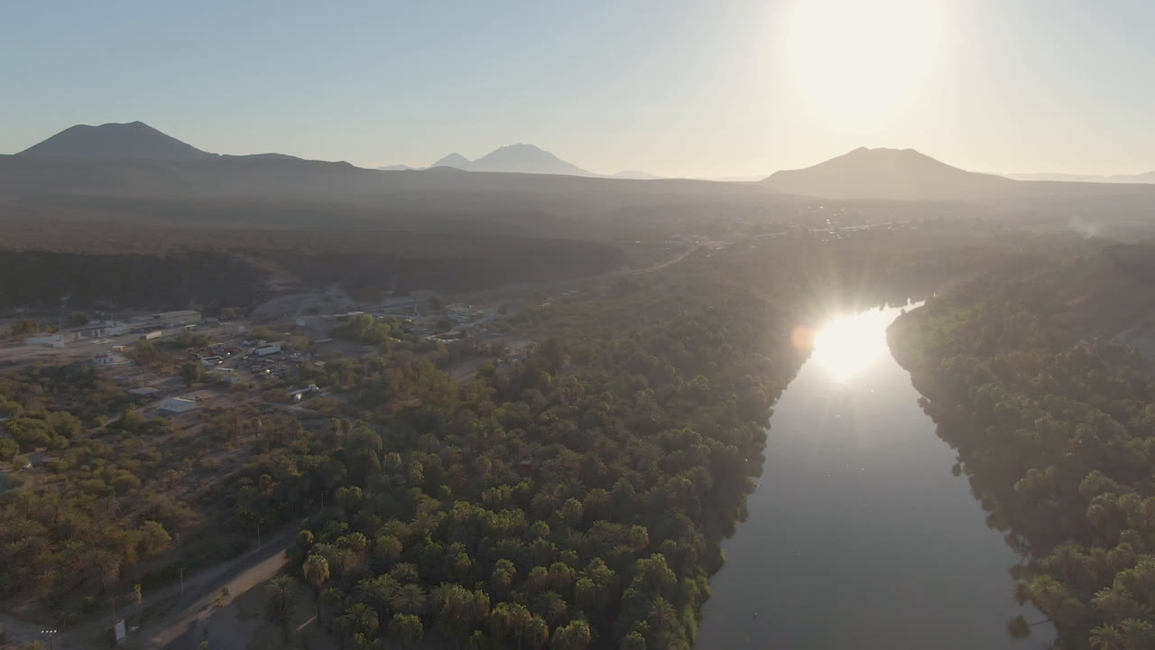 Aerial frame over San Ignacio, Baja California Sur, showing a reflective river below bright sun glare, dense trees on both banks, hazy mountain silhouettes, and no visible people.