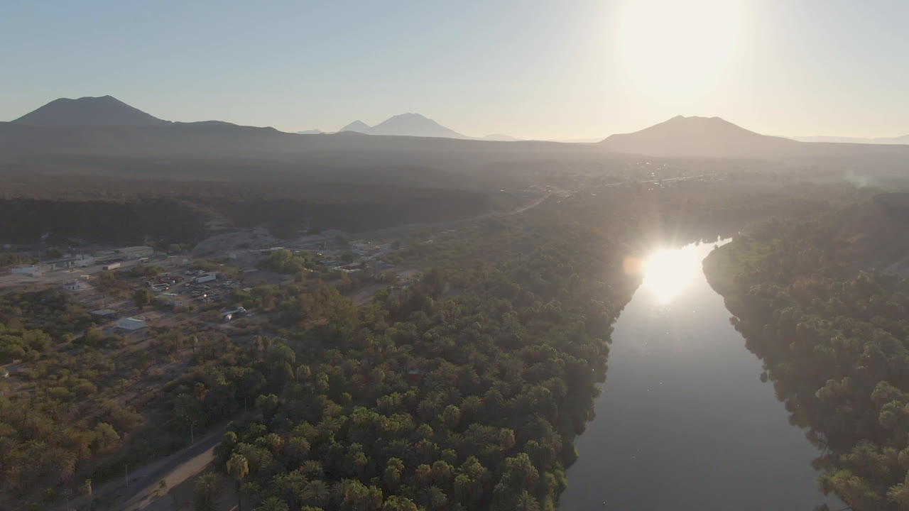 Aerial view in San Ignacio, Baja California Sur, with the river centered under a bright sun reflection, trees filling the lower frame, low buildings at left, and no visible people.
