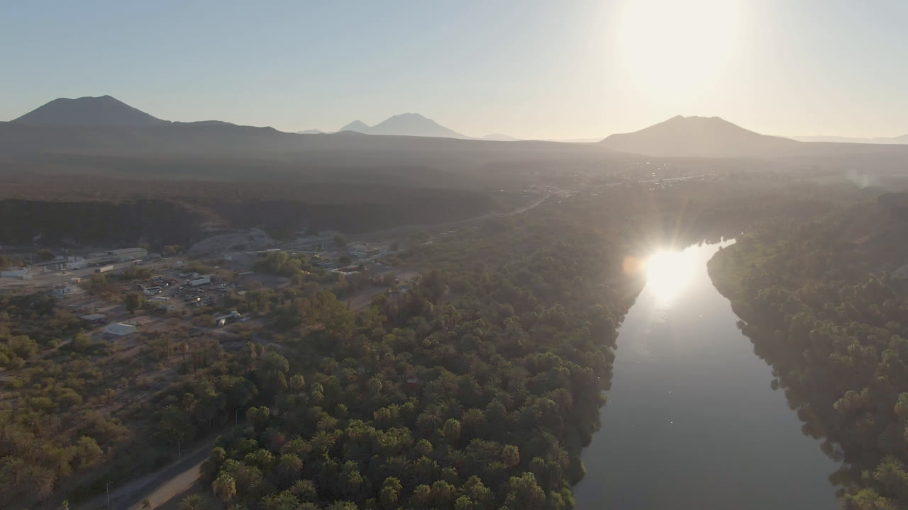Wide aerial landscape with river, green valley, small buildings, hazy mountains, and bright sky.