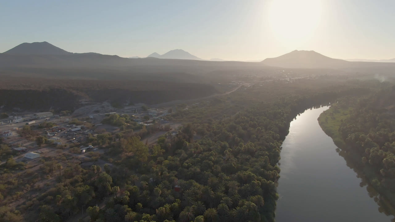 Opening aerial view over San Ignacio, Baja California Sur, with a curving river beside dense palms, low buildings at left, hazy mountains on the horizon, and no visible people.