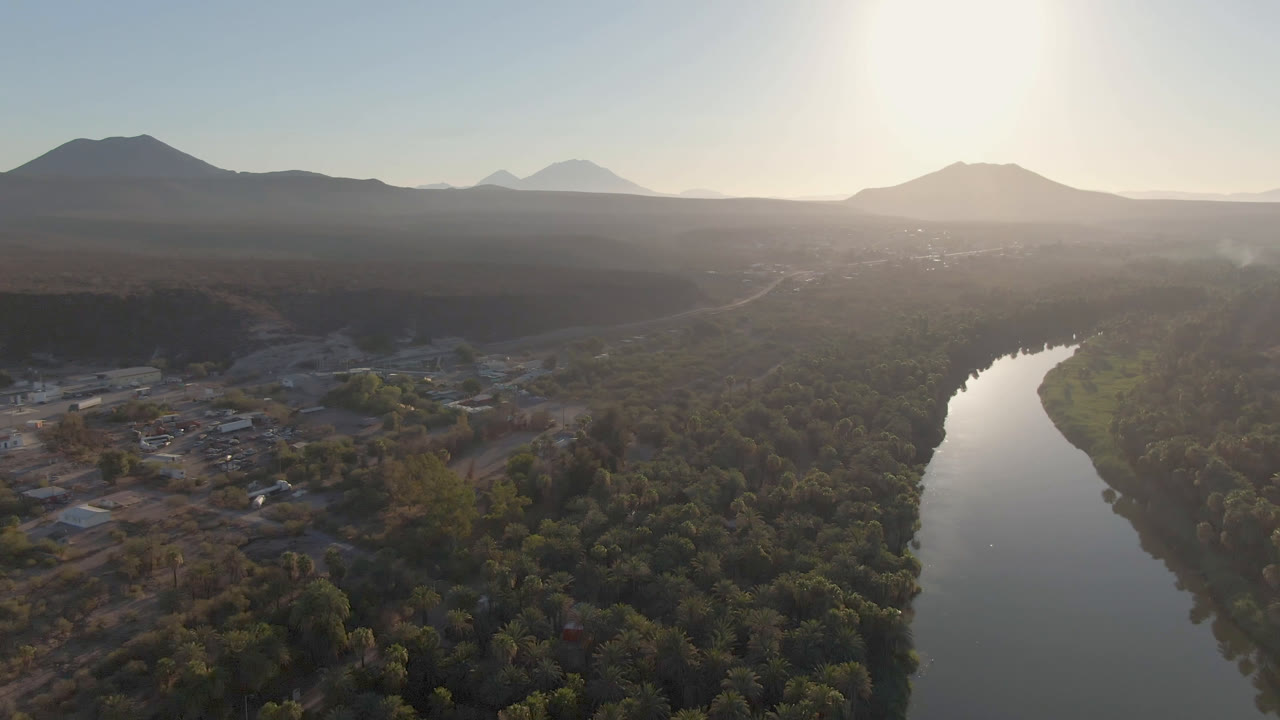Aerial frame over San Ignacio with the river bend centered lower in the view, palm canopy filling the valley floor, small structures to the left, distant mountains, bright haze, and no visible people.