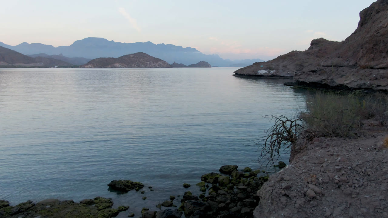 At 0:01 in Danzante Bay, the rocky point sits near the center-right of the frame, with blue water spread across the foreground, hazy mountains in back, and no visible people.