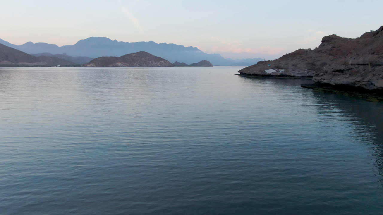 The second frame holds on a rocky island edge over blue bay water in Loreto, Mexico, with layered mountains in the distance and no visible people.