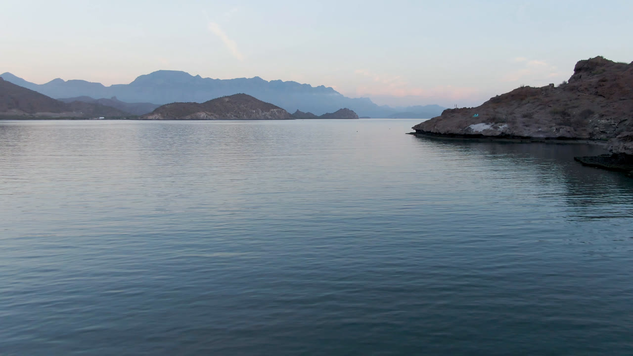 Wide view of calm coastal water in Loreto, Mexico, with a rocky island right of center, layered mountains in the distance, pale sky color, and no visible people.
