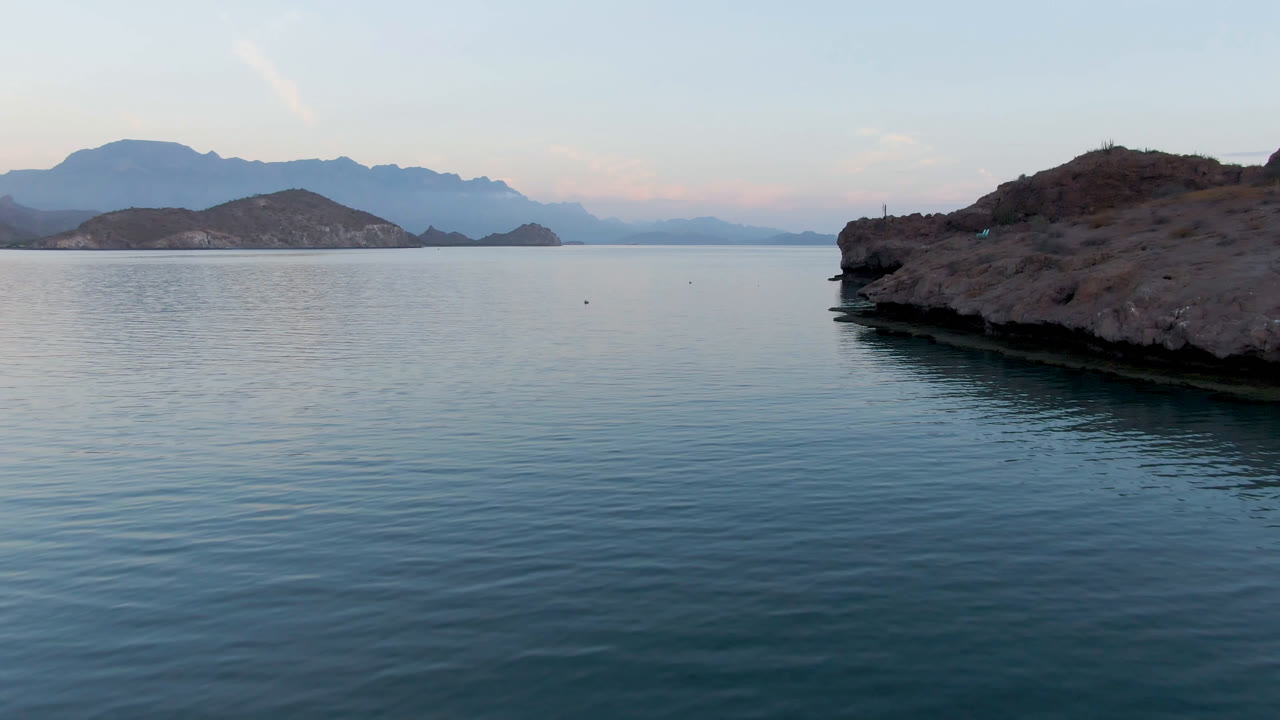Opening frame of Danzante Bay in Loreto, Baja California Sur, with calm blue water, a rocky point at right, layered mountains on the horizon, pale sky, and no visible people.