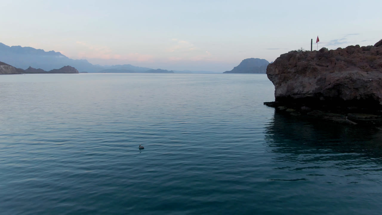 Wide view over calm water in Danzante Bay, Loreto, with a rocky point at the right edge, low island silhouettes on the horizon, pastel sky, and no visible people.