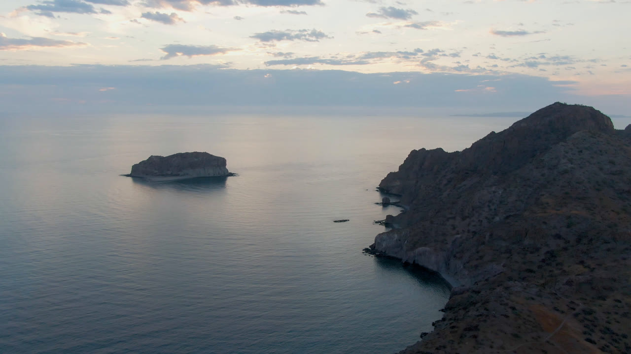Opening frame of Danzante Bay in Loreto, Mexico, with dark rocky cliffs on the right, a small island offshore on the left, calm water, pale sunrise clouds, and no visible people.
