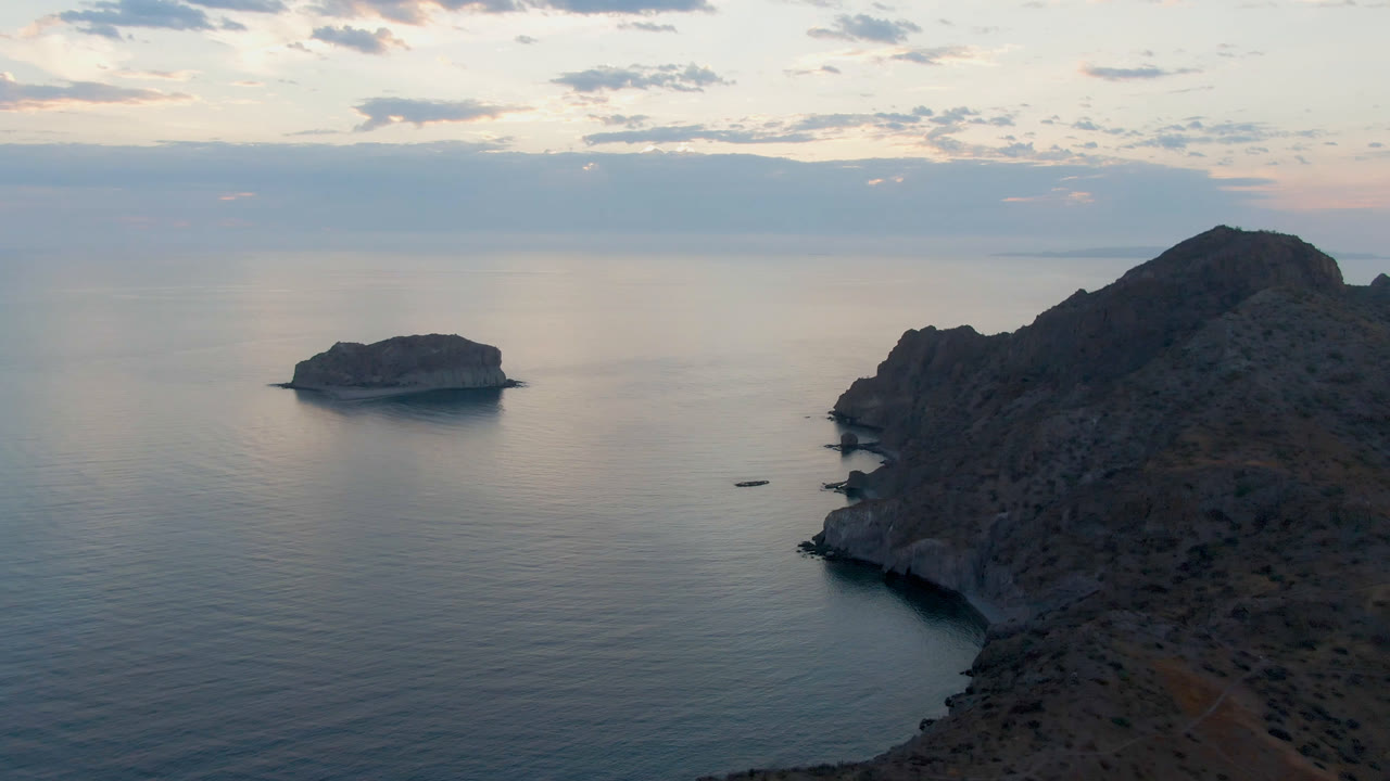 One second into the Danzante Bay clip, the rocky headland remains on the right while the small island sits across calm reflective water under layered clouds, with no visible people.
