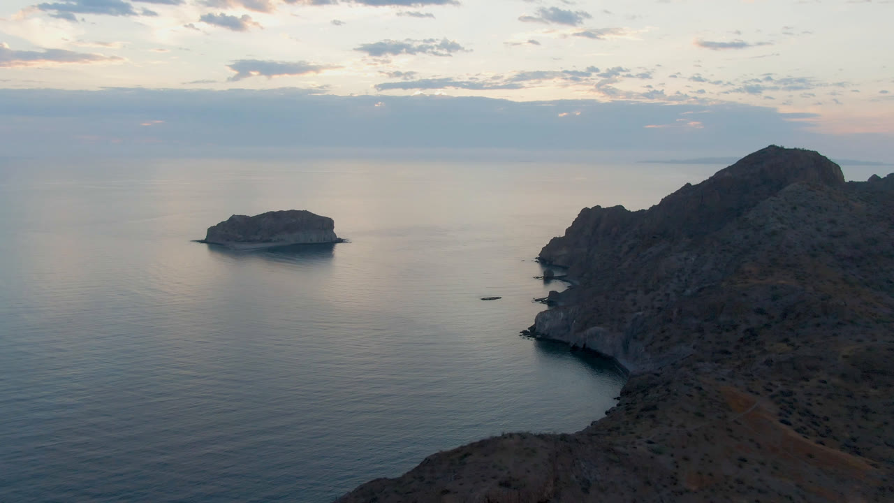 Danzante Bay, Loreto at dusk with a small boat on calm water, a rocky cliff on the right, a low island offshore, cloud cover above, and no visible people.