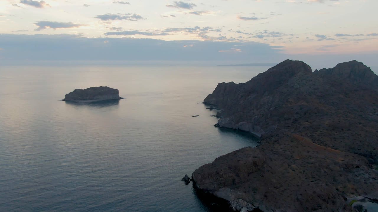 Opening frame over Danzante Bay in Loreto, Baja California Sur, with a small island on the left, rocky coast on the right, soft clouds above the horizon, and no visible people.