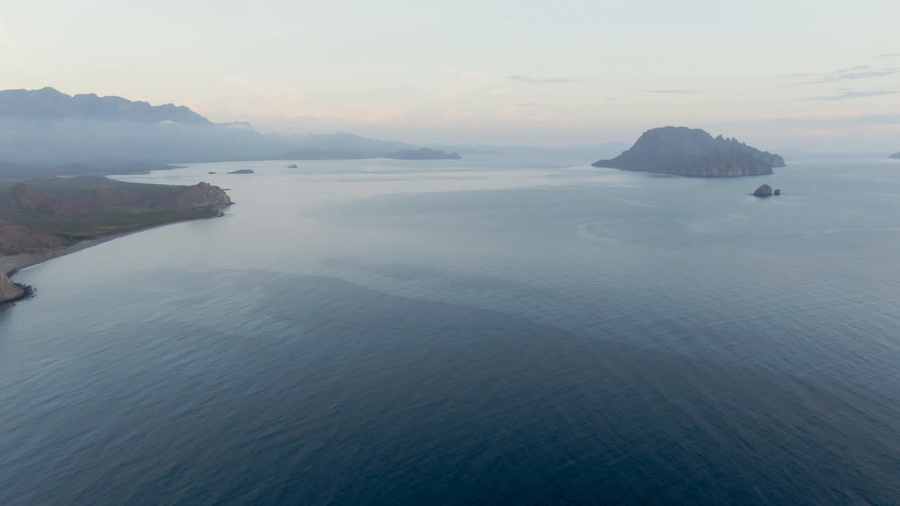 Wide aerial frame over Danzante Bay in Loreto, Baja California Sur, with blue water in front, rocky shore at left, islands to the right, hazy mountains beyond, and no visible people.