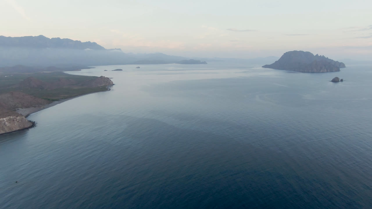 Opening aerial frame over Danzante Bay in Loreto, Baja California Sur, with blue-green water in the foreground, rocky shoreline on the left, islands to the right, hazy mountains in back, and no visible people.