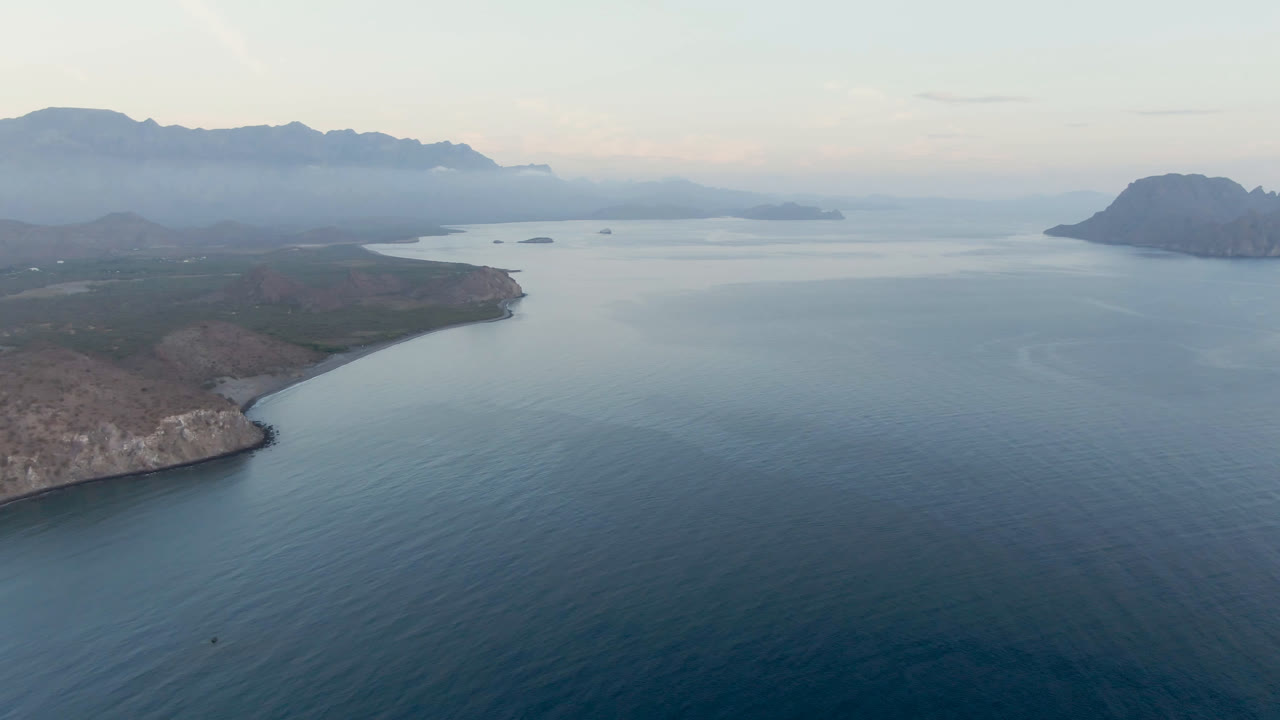 Opening aerial frame over Danzante Bay Loreto, with blue water in the foreground, a rocky shore at left, hazy mountains on the horizon, and no visible people.