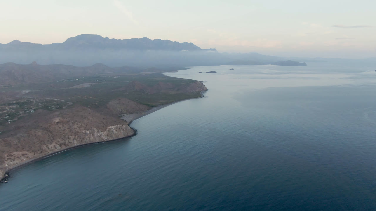 Aerial view of Danzante Bay with the rocky point lower left, broad blue water across the frame, distant island shapes, mountain ridges, and no visible people.