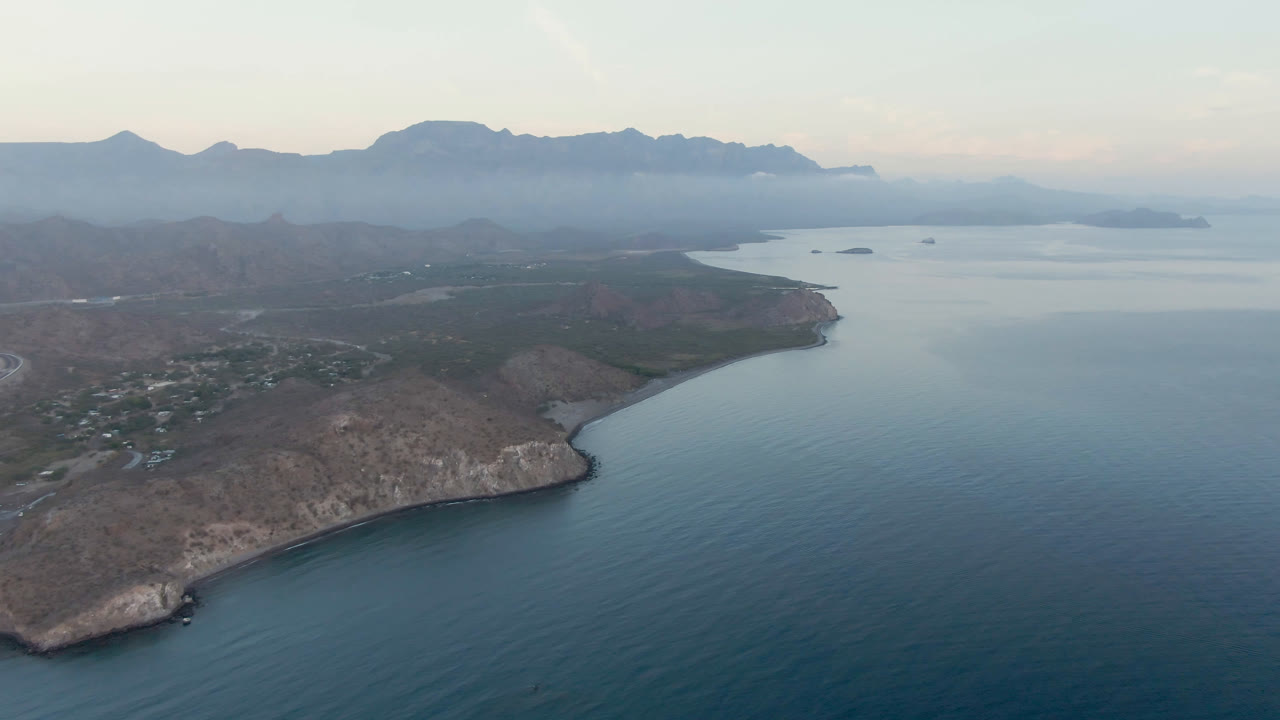 Aerial opening frame over Danzante Bay in Loreto, Baja California Sur, with blue-green water, a rocky peninsula, small buildings, distant islands, hazy mountains, and no visible people.