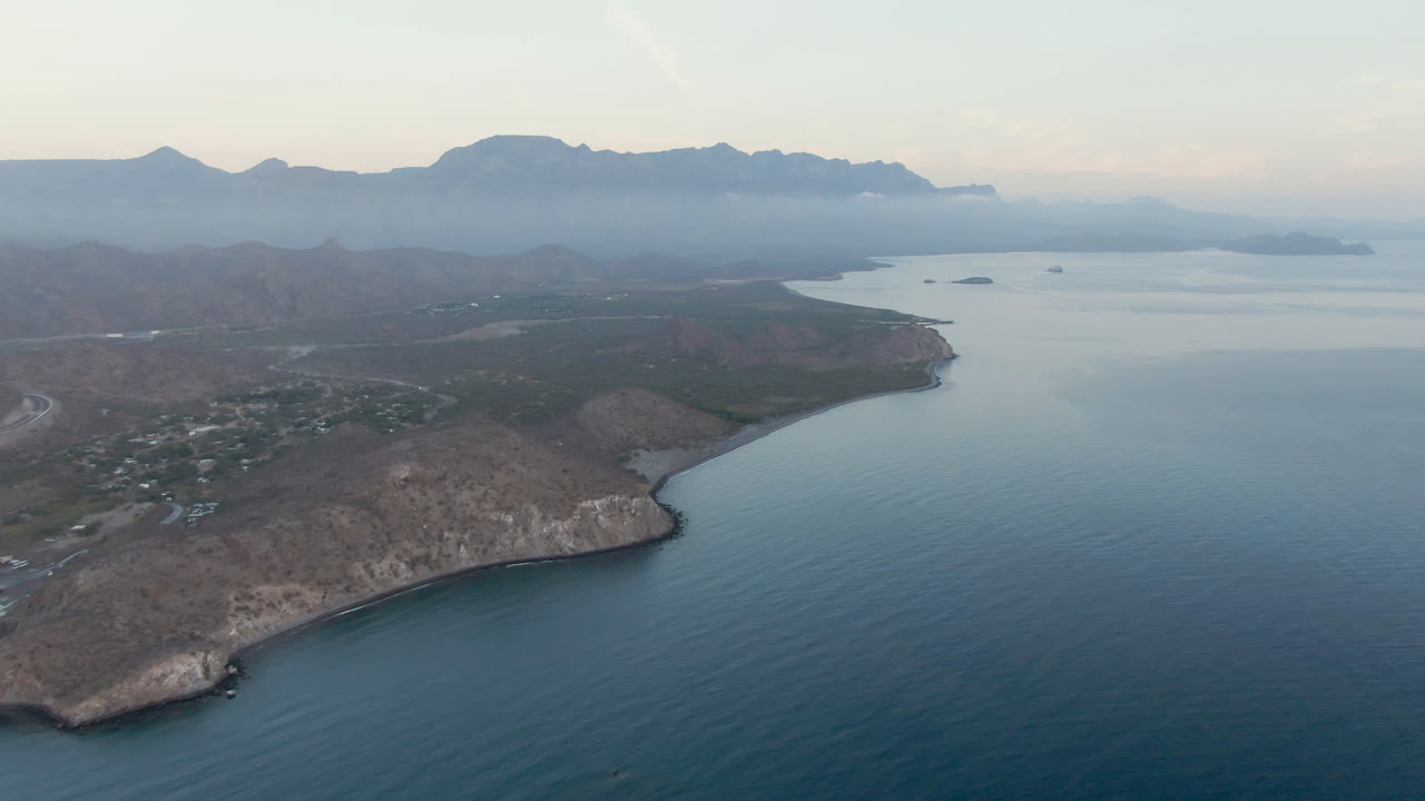 Drone frame over Danzante Bay showing the curved rocky shoreline more centrally, calm water to the right, scattered small structures inland, hazy mountain ridges, and no visible people.