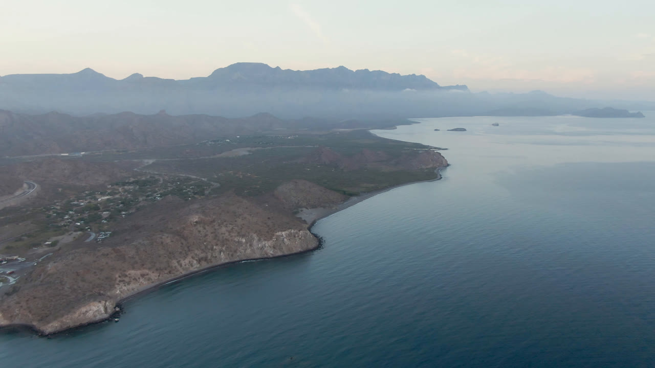 Wide aerial of coastal point, blue water, islands, and mountain backdrop